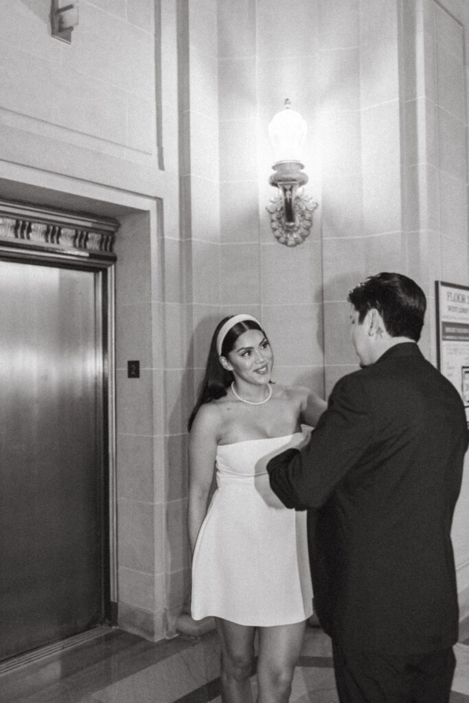 A monochrome photo depicting a woman in a stylish strapless white dress and headband, smiling at a man in a dark suit. They stand inside San Francisco City Hall near a vintage wall lamp