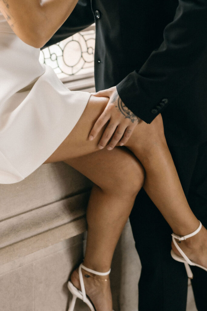 A close-up of a couple's lower bodies showing romantic intimacy during their San Francisco City Hall elopement; the man in black pants holds the calf of the woman in a white dress and heels