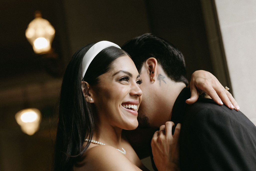 A joyful bride with a headband and pearl necklace smiling brightly as she embraces a groom, who is seen from behind with tattoos on his neck, under soft lighting at their San Francisco City Hall elopement