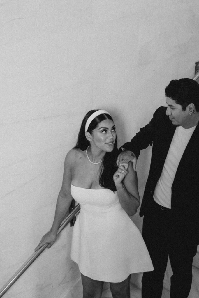 A black and white photo of a joyful woman in a white dress and headband holding hands with a man in a dark suit, as they both smile and interact on the staircase of San Francisco City Hall