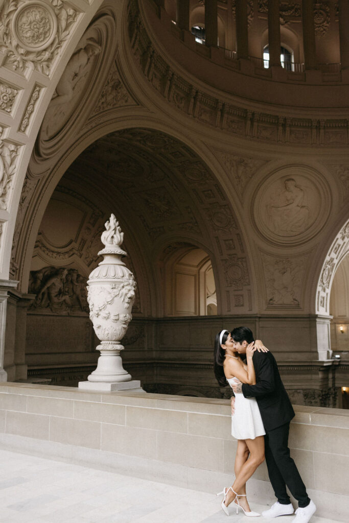 A couple kisses passionately in San Francisco City Hall with ornate architecture, intricate sculptures, and a large decorative urn, surrounded by a soft, luminous ambiance.