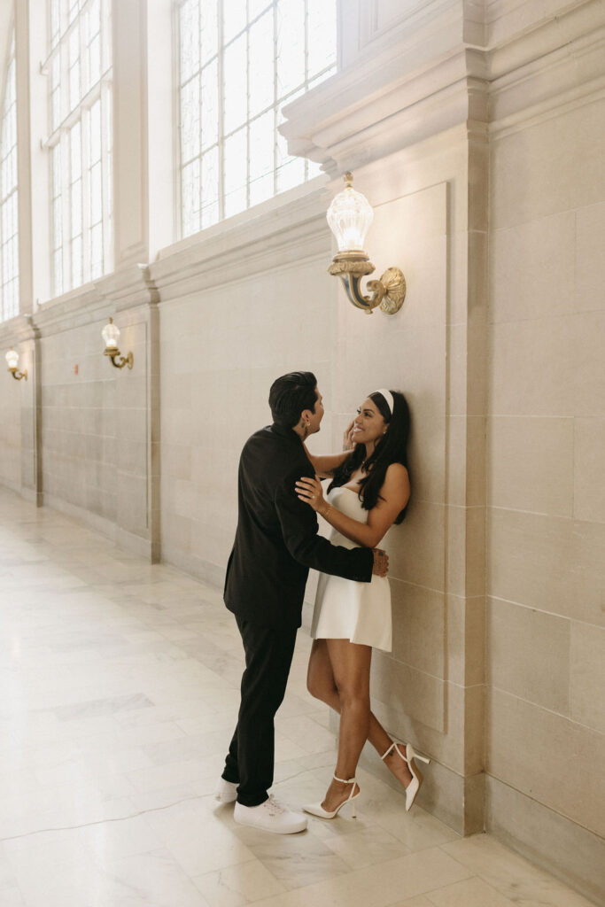 A couple in elegant attire shares a tender moment by a wall in San Francisco City Hall during their elopement, with large windows and ornate light fixtures. She wears a white dress and heels;