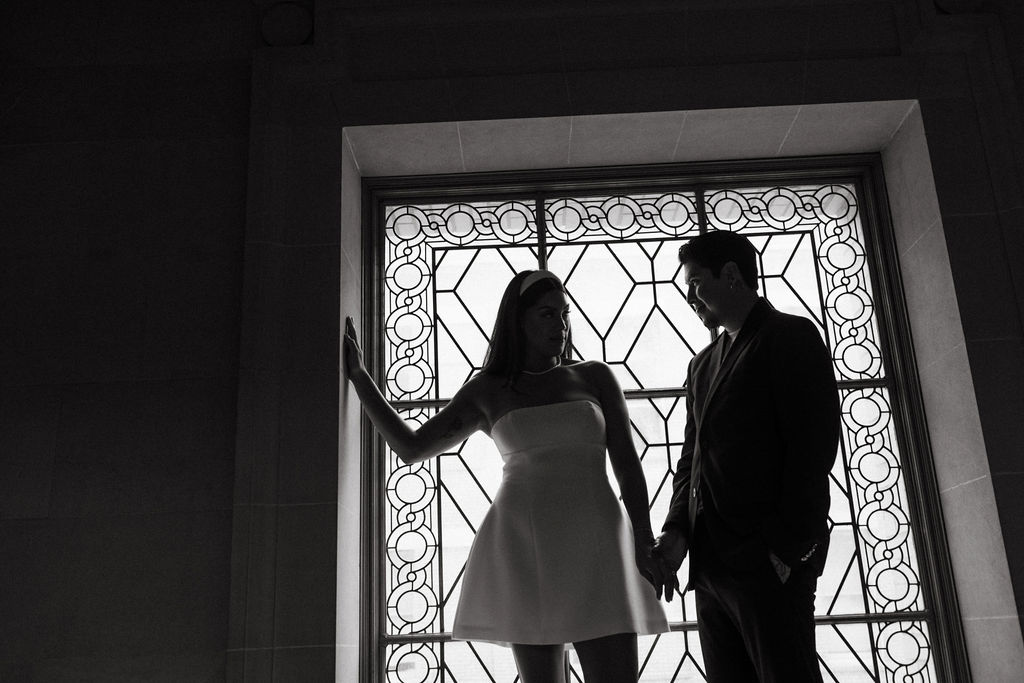 Silhouetted couple standing by an ornate stained glass window in San Francisco City Hall, engaged in a conversation. The woman gestures with her hand, casting a shadow across the man's face.