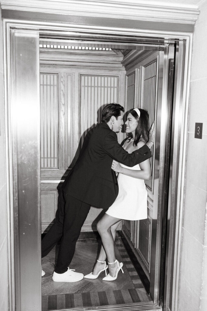 A black and white photo capturing a man in a suit and a woman in a dress during their San Francisco City Hall elopement, having a playful moment inside an elevator. 