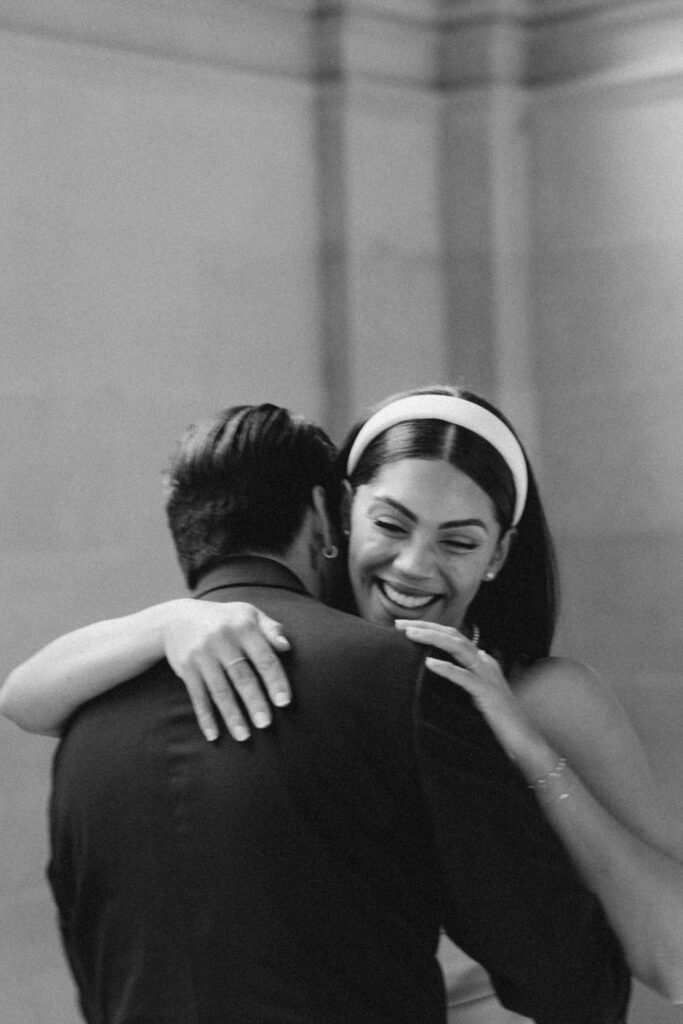 A black and white photo of a woman smiling joyfully during her San Francisco City Hall elopement, embracing a man while focusing on her expression as she looks over his shoulder.