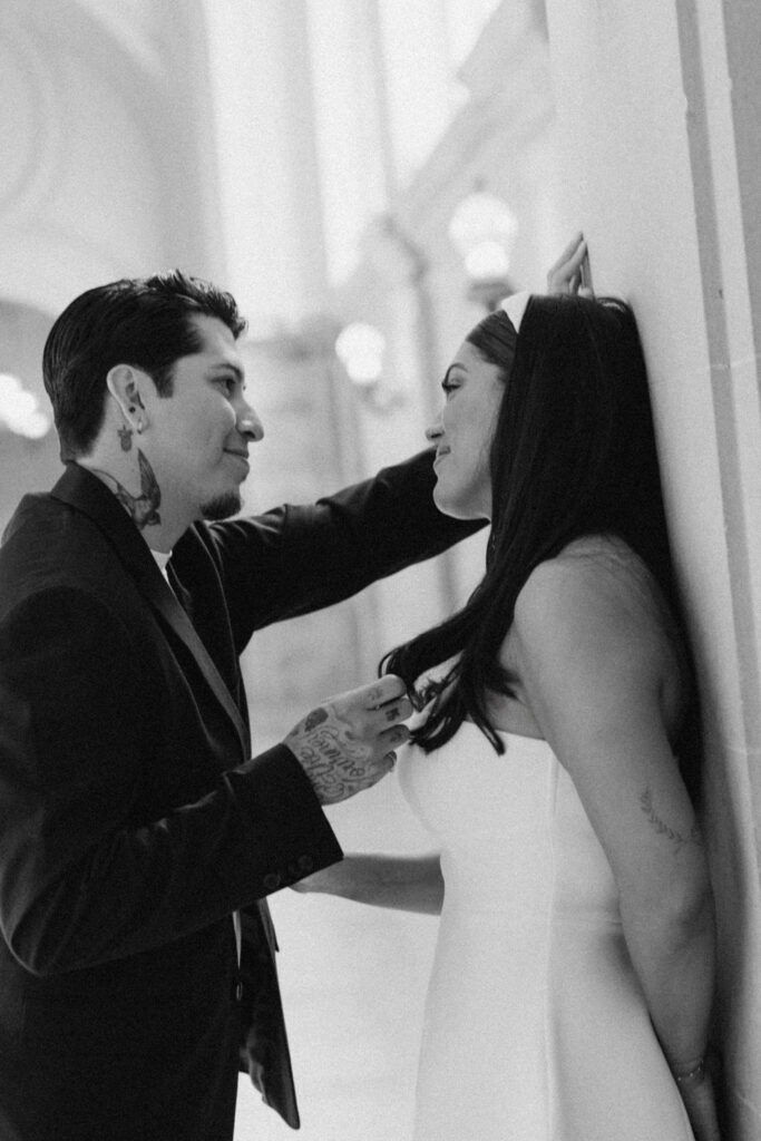 A black and white image of a man tenderly touching a woman's face as they stand close together in a columned corridor at San Francisco City Hall, expressing affection and intimacy during their elopement